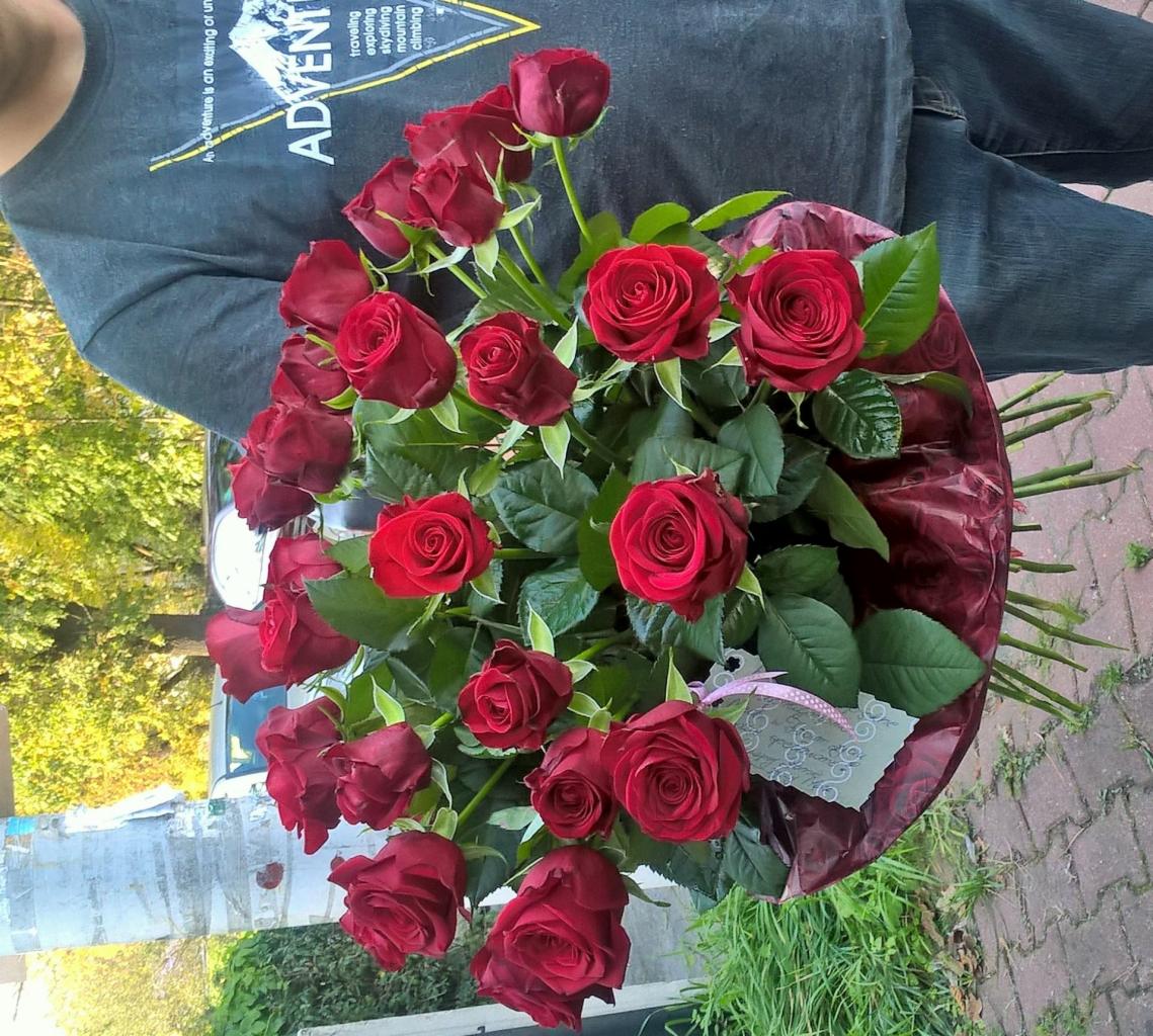 Mężczyzna trzyma bukiet czerwonych róż w bordowym opakowaniu z delikatną wstążką i kartką z życzeniami, na tle chodnika i zieleni.