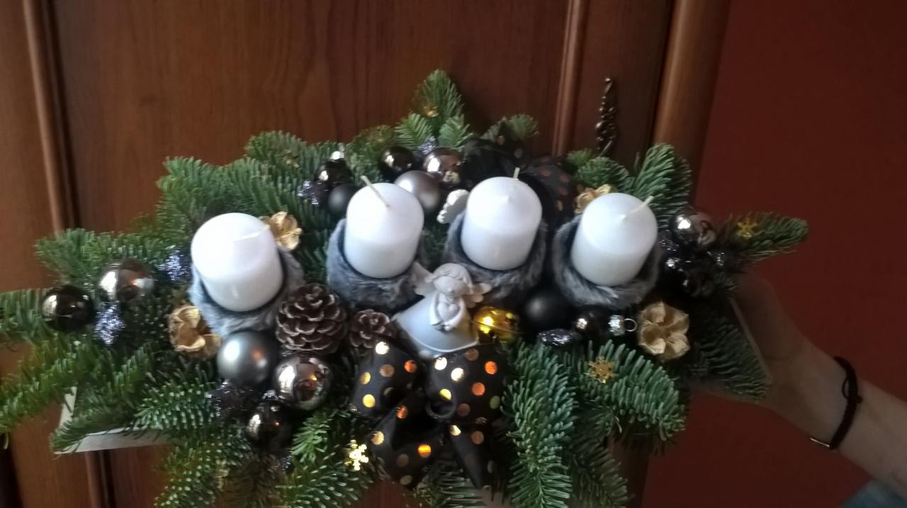 Dekoracja adwentowa z czterema białymi świecami w futerkowych otoczkach, srebrnymi bombkami, szyszkami, czarną kokardą w złote kropki i figurką anioła, na tle drewnianych drzwi i czerwonej ściany.