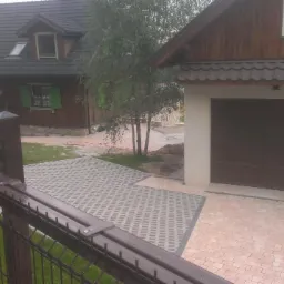 Widok na posesję z brązową bramą garażową, domkiem w stylu rustykalnym, ażurową kostką brukową i fragmentem metalowego ogrodzenia z lampą ostrzegawczą.