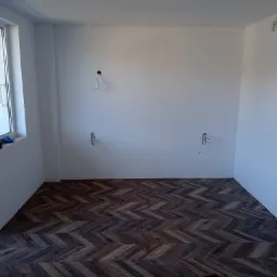 Ułożona podłoga z paneli w jodełkę w pustym, świeżo pomalowanym na biało pokoju z wystającymi przewodami elektrycznymi i oknem balkonowym.