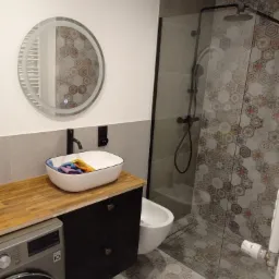 Nowoczesna łazienka z drewnianym blatem, okrągłym lustrem z podświetleniem LED, kabiną prysznicową z czarnymi okuciami i mozaiką heksagonalną na ścianach oraz pralką w zabudowie.