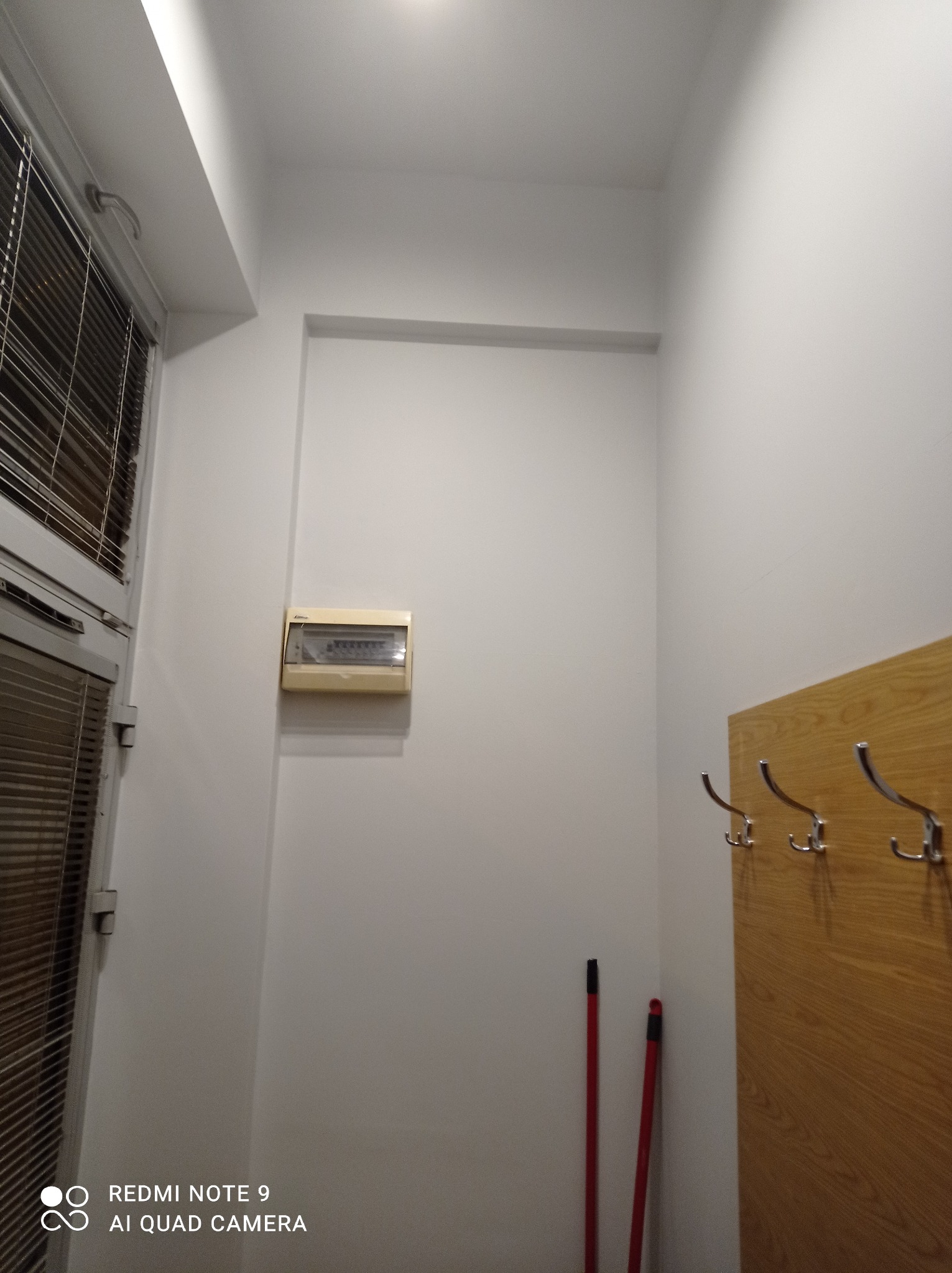 Wnętrze korytarza po odświeżeniu: białe ściany, drewniany panel z wieszakami, skrzynka elektryczna pod sufitem oraz dwie czerwone rurki w rogu.