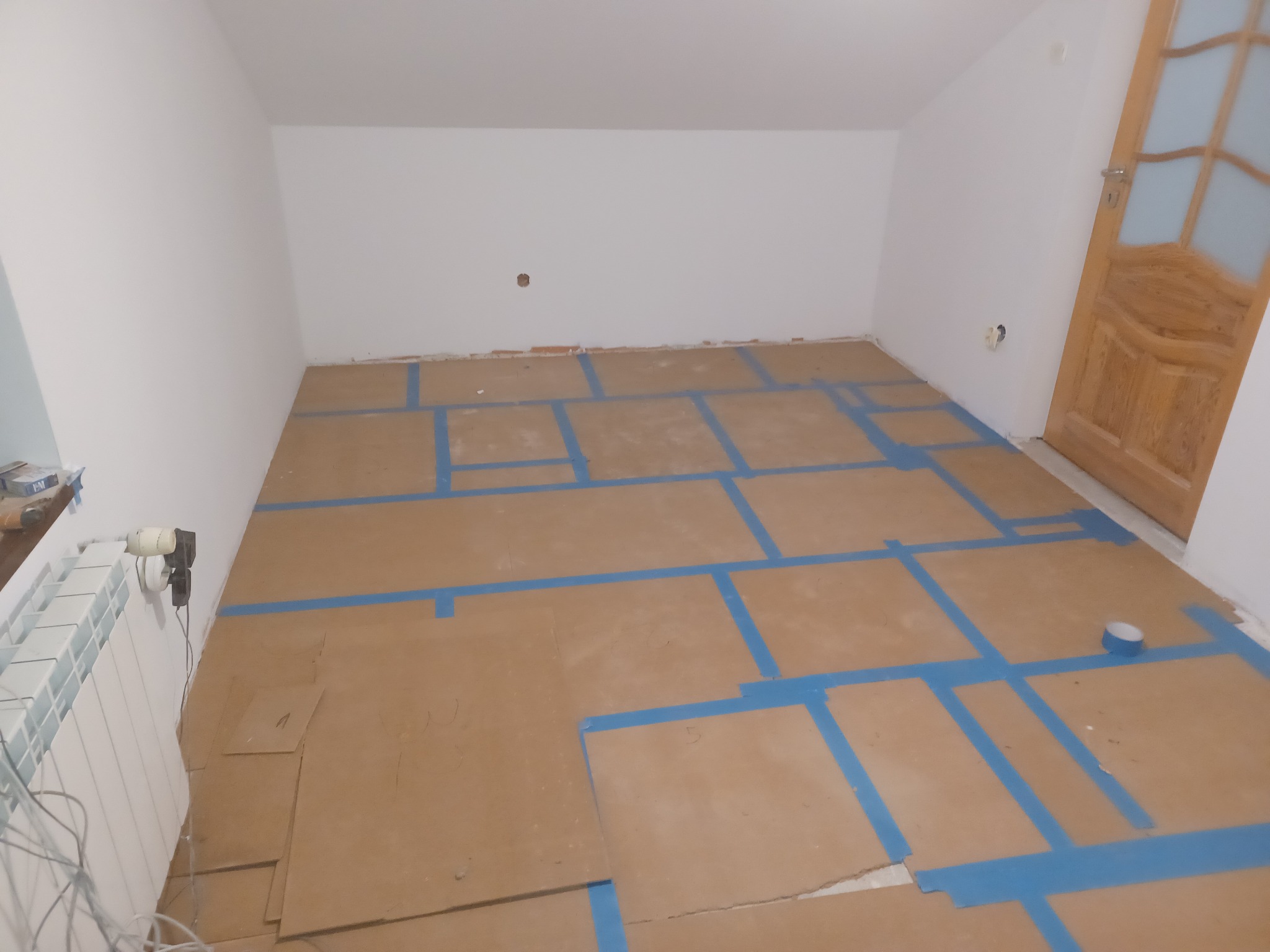 Przygotowanie podłoża pod panele: rozłożone płyty podkładowe z zaznaczonymi niebieską taśmą polami na podłodze w pokoju z grzejnikiem i drewnianymi drzwiami.