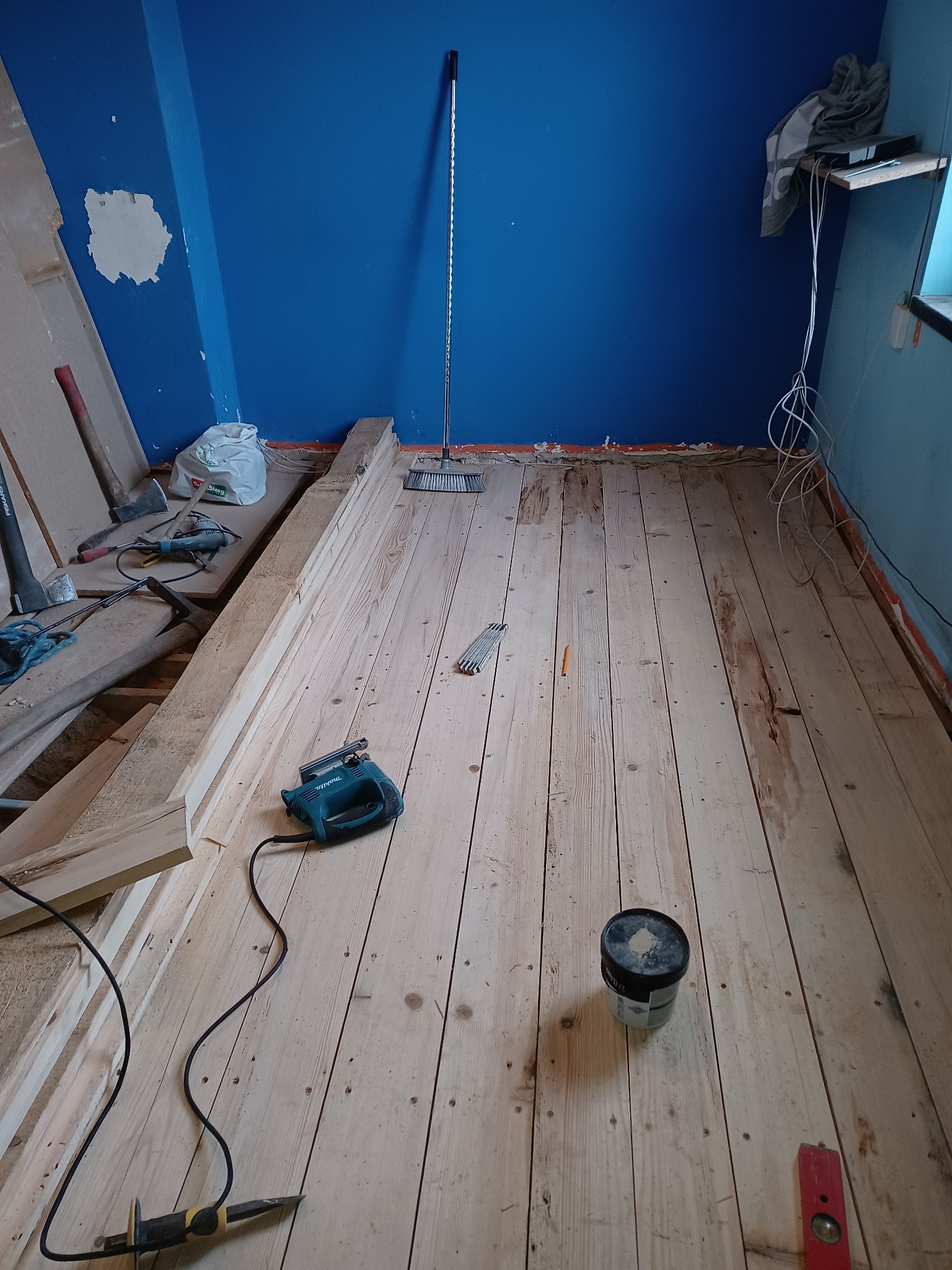 Surowa podłoga z desek w trakcie montażu, widoczna wyrzynarka, poziomica i puszka wypełniacza do drewna, ściana pomalowana na niebiesko z widocznymi śladami po usuniętej farbie.