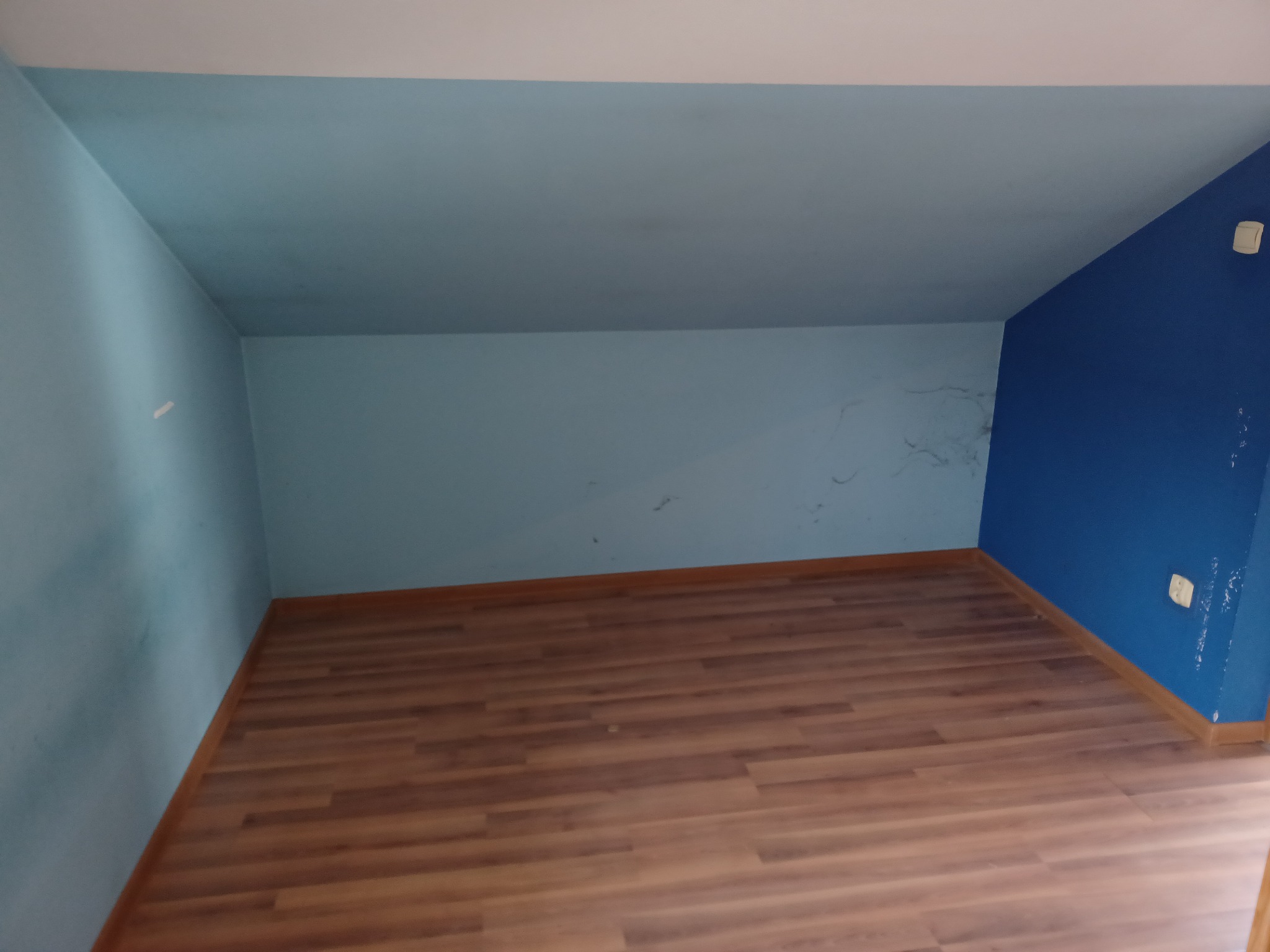 Poddasze po remoncie z drewnopodobną podłogą, ściany pomalowane na jasny i ciemny odcień niebieskiego, widoczne ślady po taśmie malarskiej i zabrudzenia na ścianach.