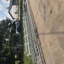 FRENO Spółka z ograniczoną odpowiedzialnością - Biała, metalowa drabina z osłoną zabezpieczającą przed upadkiem, leżąca poziomo na betonowej powierzchni, z budynkami i zielenią w tle.