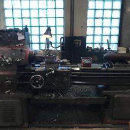 FRENO Spółka z ograniczoną odpowiedzialnością - Stary, używany tokarka w warsztacie, z widocznymi wiórami metalowymi i lampą oświetlającą obszar roboczy, na tle okna z luksferami.