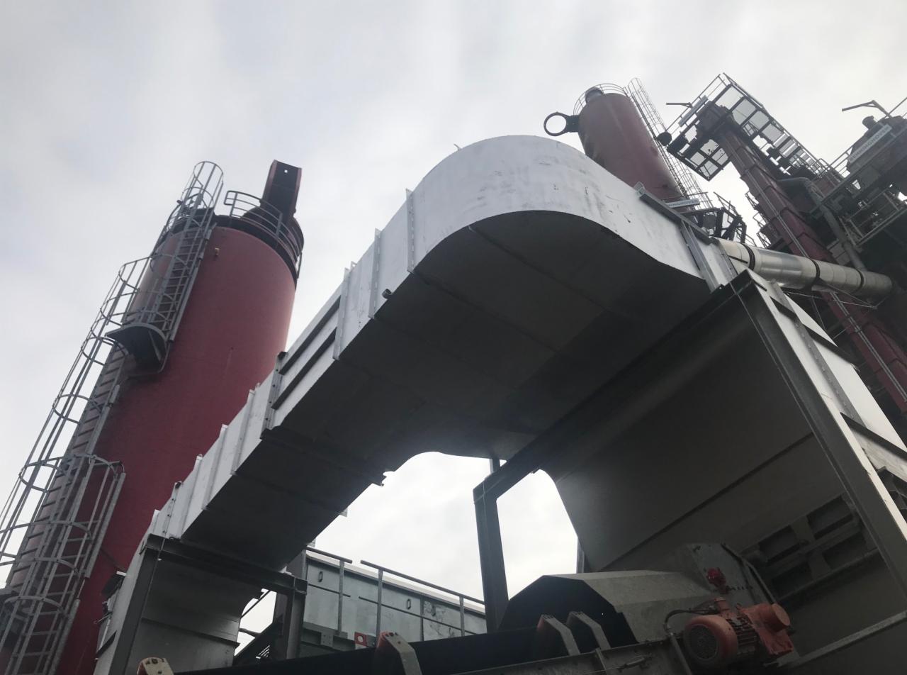 Widok z dołu na konstrukcję stalową hali z czerwonymi kominami i elementami transportowymi pod pochmurnym niebem.