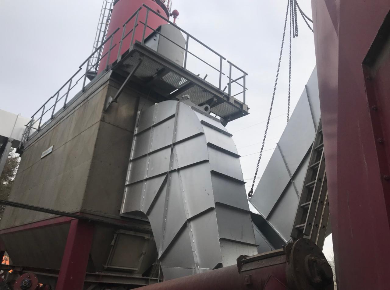 Montaż metalowej konstrukcji hali z czerwonym elementem na szczycie, widoczne łańcuchy podtrzymujące element, pochmurne niebo w tle.