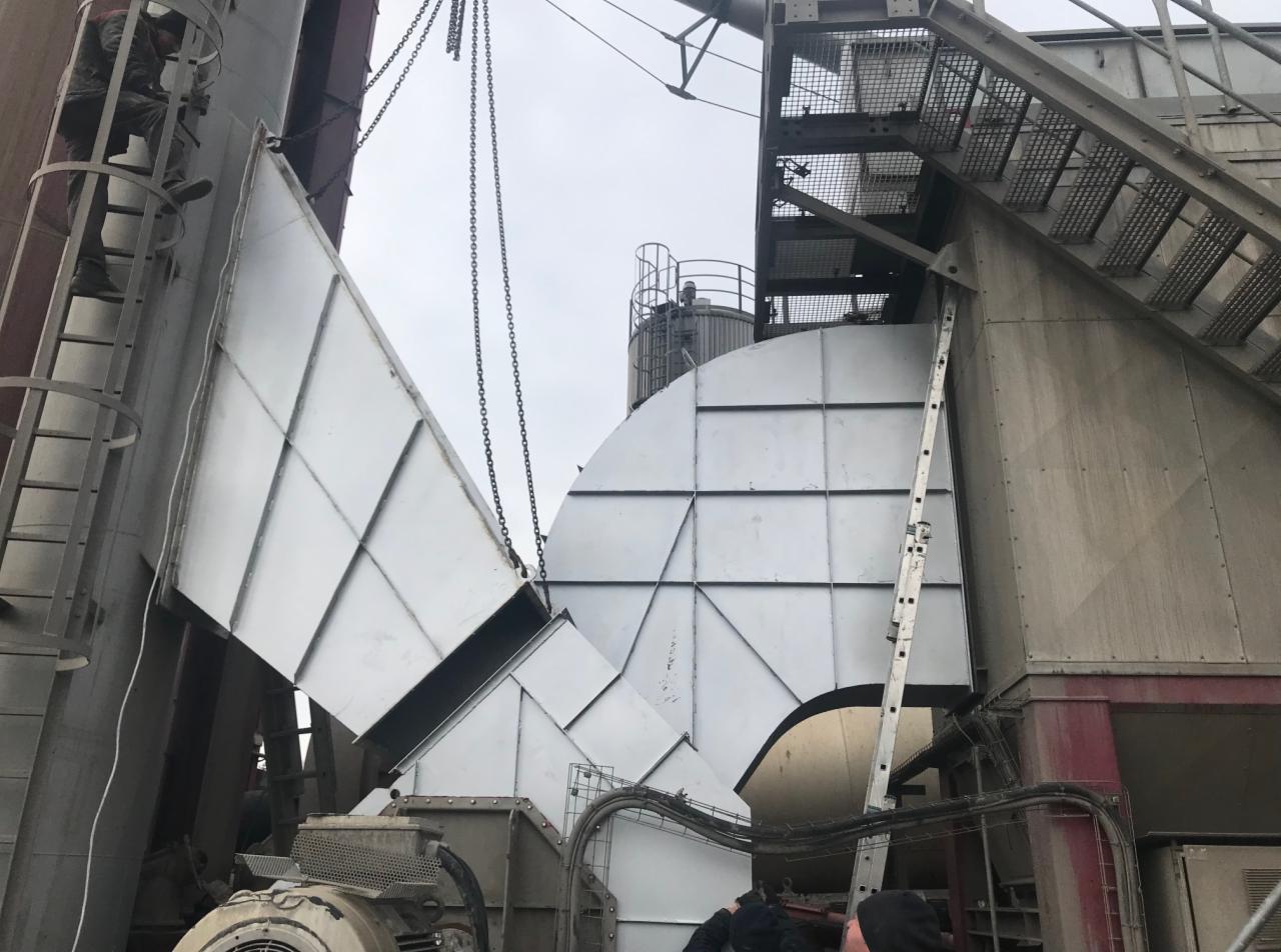 Montaż segmentów konstrukcji stalowej hali przemysłowej z użyciem łańcuchów i dźwigu, widoczne drabiny i elementy instalacji.