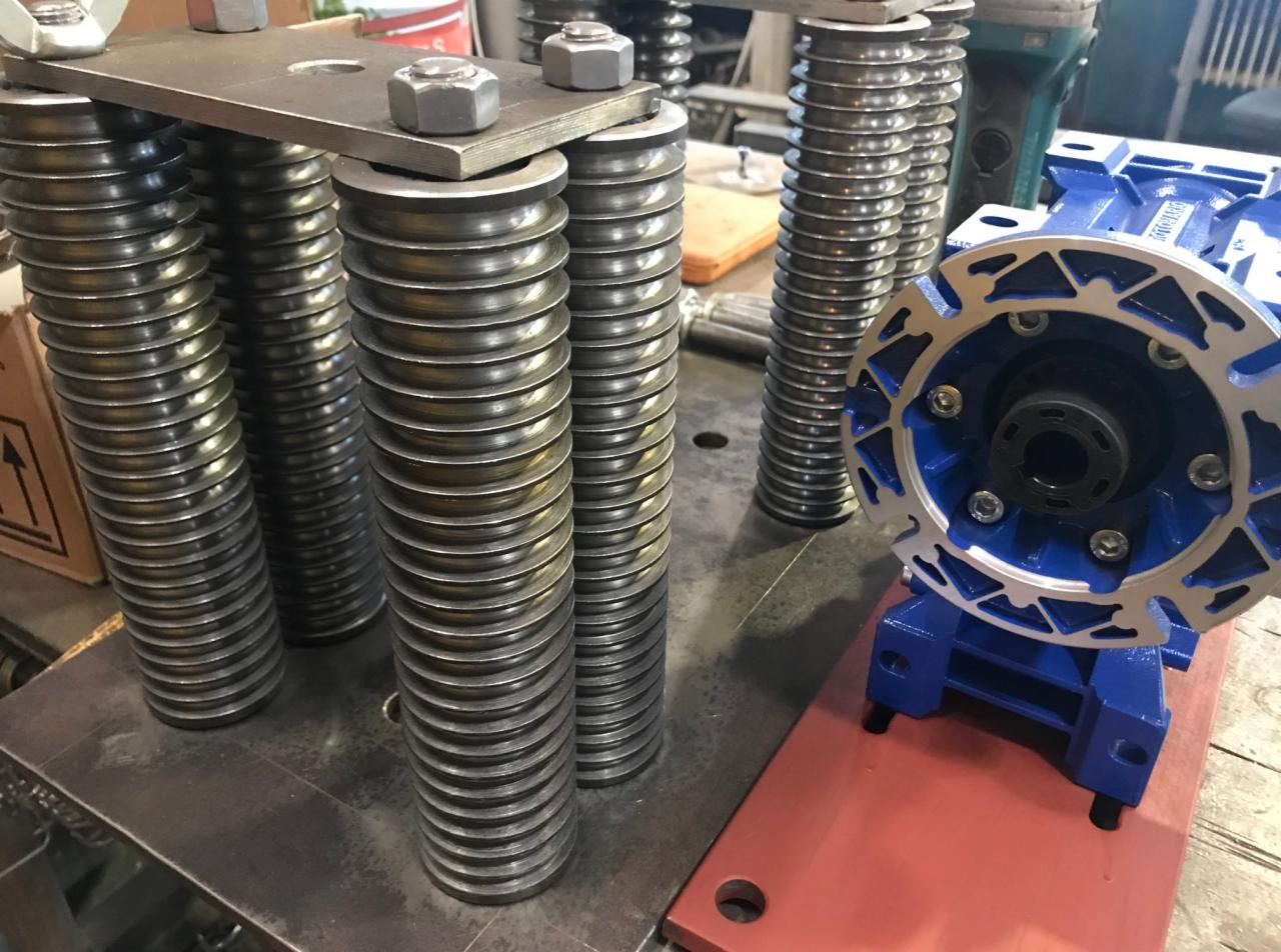 Metalowe kolumny sprężynowe połączone płytą z nakrętkami, obok niebieska przekładnia ślimakowa na czerwonej podstawie.