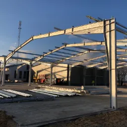 Wznoszenie szkieletu hali stalowej w słoneczny dzień, widoczne dźwigi i przygotowane elementy konstrukcyjne.