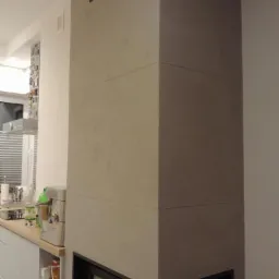 Obudowa kominka wykończona płytami z szarego betonu architektonicznego z czarnymi detalami, widok z boku na tle kuchni.