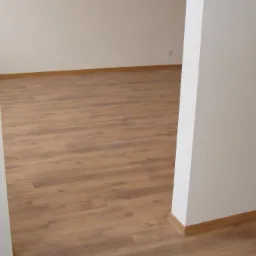 Jasne panele podłogowe ułożone w pustym pokoju, widoczne listwy przypodłogowe z imitacji drewna i fragment ściany z obrazkiem