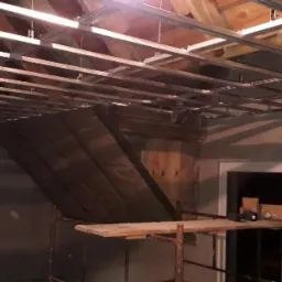 Montaż metalowej konstrukcji sufitu podwieszanego w pokoju na poddaszu, widoczne krokwie drewniane, fragment ściany z oknem i rusztowanie z narzędziami.