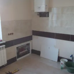 Remont łazienki w Tucholi: Widok na częściowo wykończoną ścianę z beżowymi płytkami, brązowym pasem dekoracyjnym, grzejnikiem, oraz instalacją centralnego ogrzewania wnęce. Na podłodze widoczne...