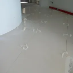 Układanie jasnych płytek podłogowych z użyciem krzyżyków dystansowych, widoczna poziomica i surowy stan wykończenia ścian w tle.