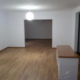 Wykończone wnętrze mieszkania z panelami podłogowymi, świeżo pomalowanymi ścianami na biało, z widocznym przejściem do kolejnego pomieszczenia i zabezpieczoną folią szafką.