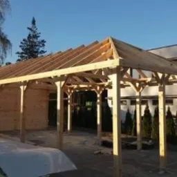 Konstrukcja drewniana z widoczną więźbą dachową w trakcie montażu, jasne drewno, błękitne niebo w tle, widoczne elementy budynku mieszkalnego.