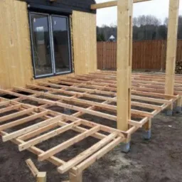 Konstrukcja drewniana tarasu w trakcie budowy, z widocznymi słupami nośnymi i legarami, na tle budynku z drewnianą elewacją i dużym oknem przesuwnym.
