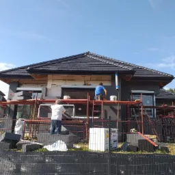 Dwóch pracowników na rusztowaniu podczas prac elewacyjnych domu jednorodzinnego. Widoczne styropianowe płyty i szara elewacja. Częściowo ocieplony budynek w słoneczny dzień.