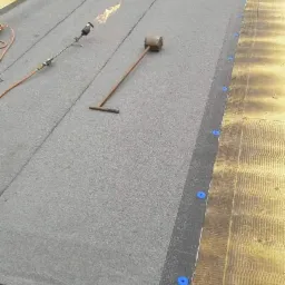 Prace dekarskie: papa termozgrzewalna układana na dachu z użyciem palnika dekarskiego i dociskana wałkiem, widoczne niebieskie kołki mocujące.