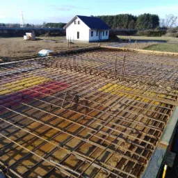 Zbrojenie płyty fundamentowej z widocznym deskowaniem i wystającymi prętami, w tle biały dom jednorodzinny w stanie surowym otwartym i błękitne niebo.