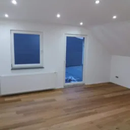 Jasne drewniane panele podłogowe w nowym pokoju z białymi ścianami, grzejnikiem i oknem z widokiem na niebieską elewację budynku.