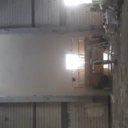 Pracownik w kamizelce odblaskowej na podnośniku koszowym przy ścianie w hali, przygotowanej do wykończenia, widoczne ślady po tynkowaniu.