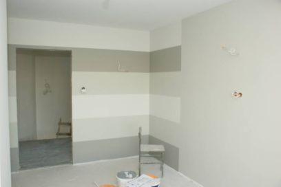 Pokój po częściowym pomalowaniu ścian w poziome pasy szarości i bieli, widoczne puszki z farbą, krzesło i niedokończone gniazdka elektryczne.