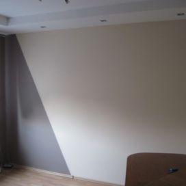 Ściana w pokoju po świeżym malowaniu z dwukolorowym wykończeniem: beżowa powierzchnia przechodzi w szary trójkąt przy krawędzi. Widoczne oświetlenie sufitowe i fragment podłogi.