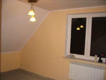 Pomalowane na ciepły, brzoskwiniowy odcień ściany poddasza w Lesznie. Widoczne okno PCV z białymi ramami, grzejnik pod oknem i lampa sufitowa z dwoma kloszami. Na parapecie leżą szpachelka i rolka...