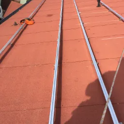 Montaż aluminiowych profili na dachu pokrytym papą termozgrzewalną, przygotowanie do instalacji paneli fotowoltaicznych, widoczny sprzęt monterski i lina zabezpieczająca.