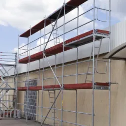 Wysokie rusztowanie modułowe z czerwonymi podestami, ustawione przy beżowej ścianie budynku z zamurowanym oknem, na brukowanym placu, z fragmentem drugiego rusztowania widocznym po lewej stronie.