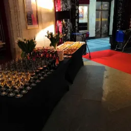 Stół z czarnym obrusem zastawiony kieliszkami z białym winem, butelkami, talerzami z przekąskami i wazonami z białymi tulipanami w eleganckim wnętrzu z czerwonym dywanem.