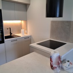 Usługi Remontowe - Nowoczesna kuchnia z jasnymi szafkami, czarną armaturą i płytą indukcyjną. Widoczny fragment blatu z akcesoriami. Minimalistyczny design, neutralna kolorystyka.