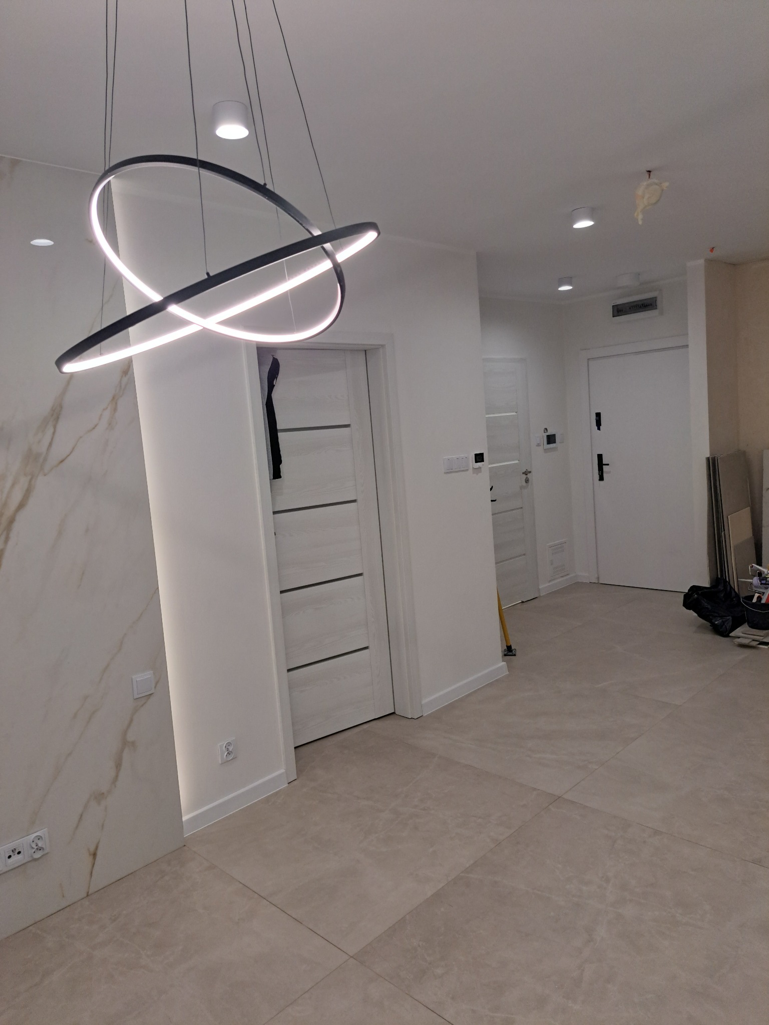 Nowoczesny przedpokój po remoncie: beżowe płytki, drzwi z poziomymi frezami, marmurowa ściana, designerska lampa. Widoczne ślady po remoncie na suficie.