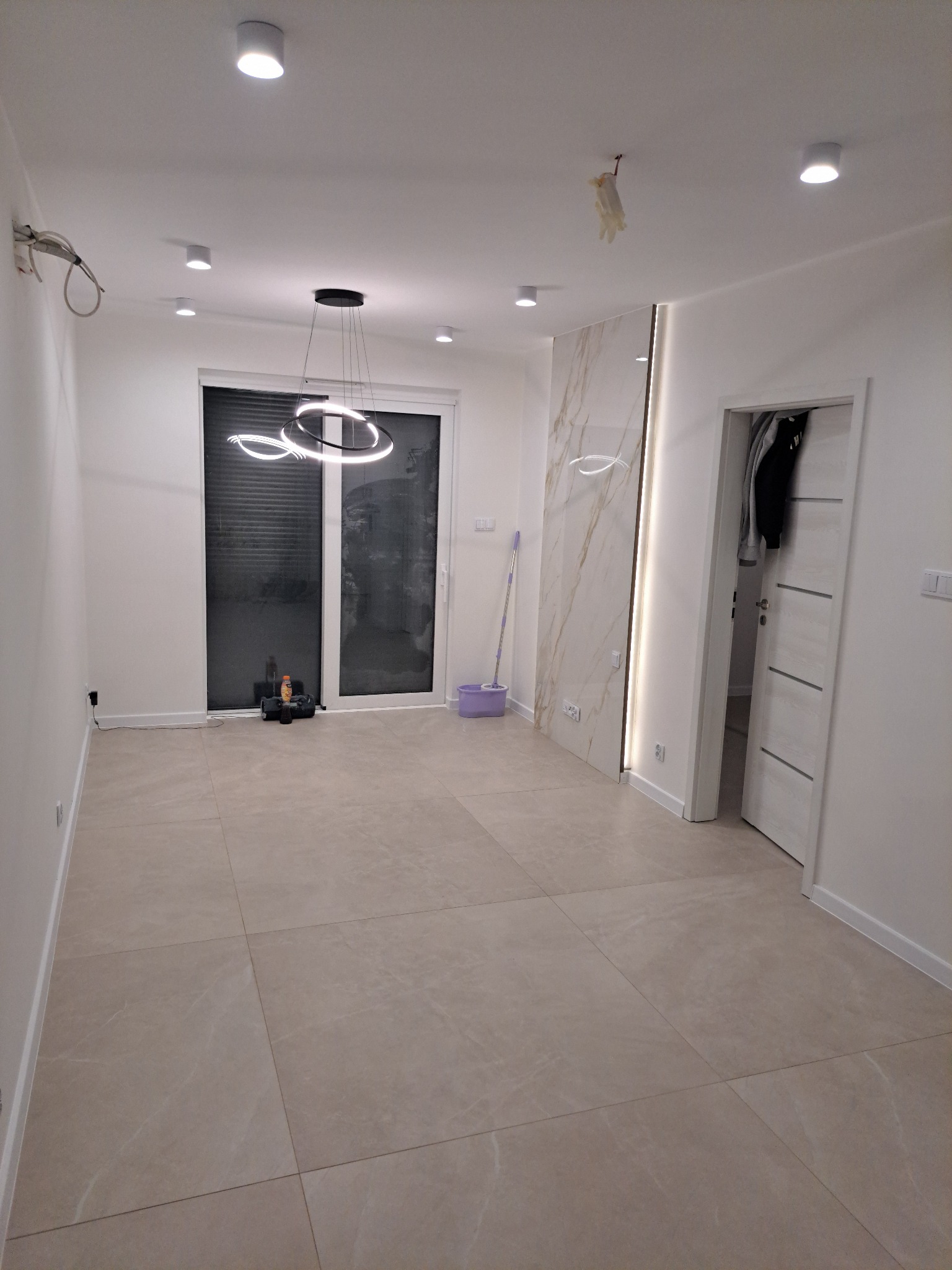 Jasne wnętrze po remoncie: beżowe płytki, białe ściany z marmurowym akcentem, nowoczesna lampa wisząca. Widoczny otwarty pokój z elementami dekoracyjnymi i mopem.