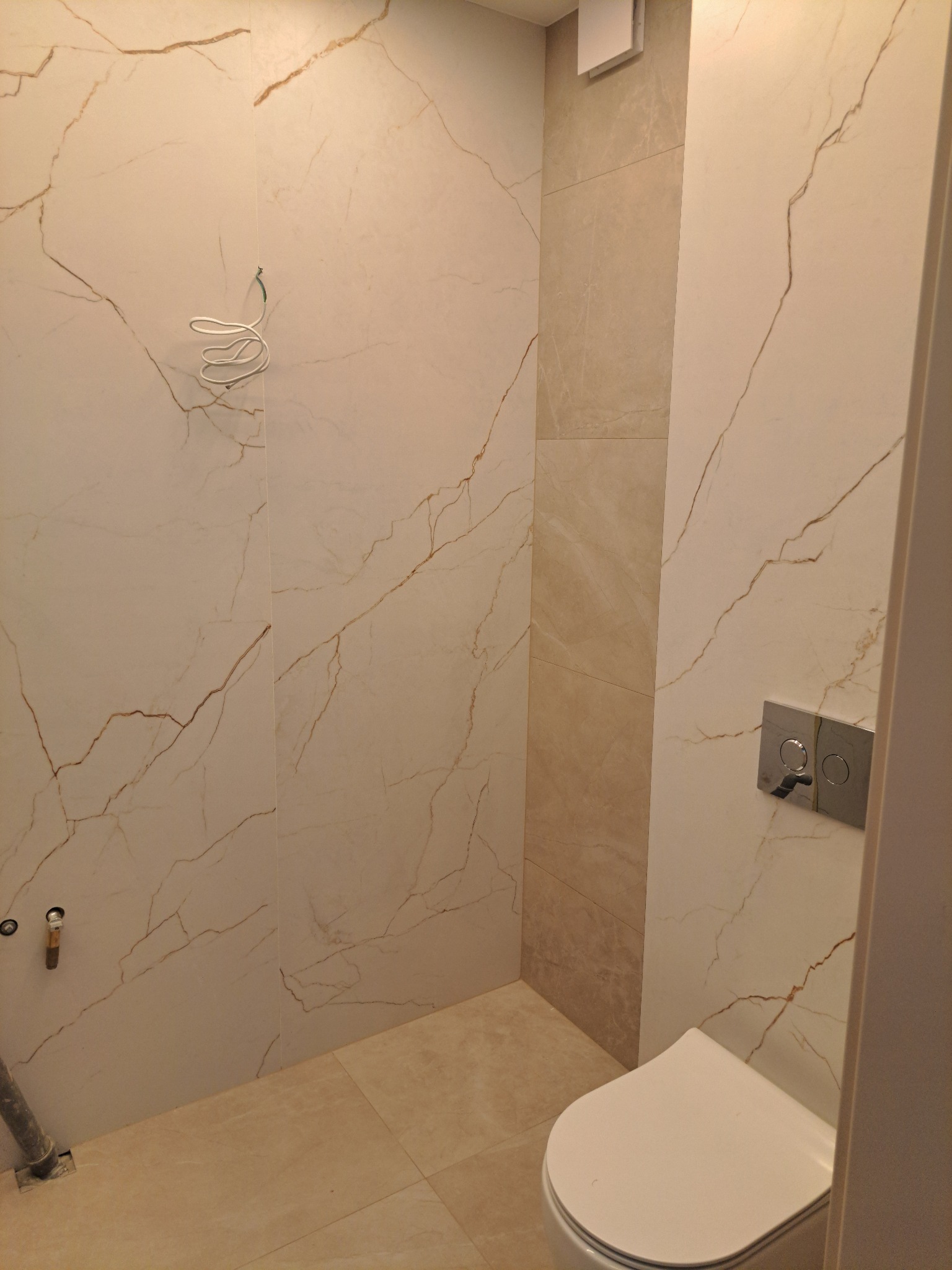 Jasna łazienka z marmurowymi płytkami, widoczne elementy instalacji, biała toaleta i fragment drzwi. Minimalistyczny design, ciepłe odcienie beżu i złota.