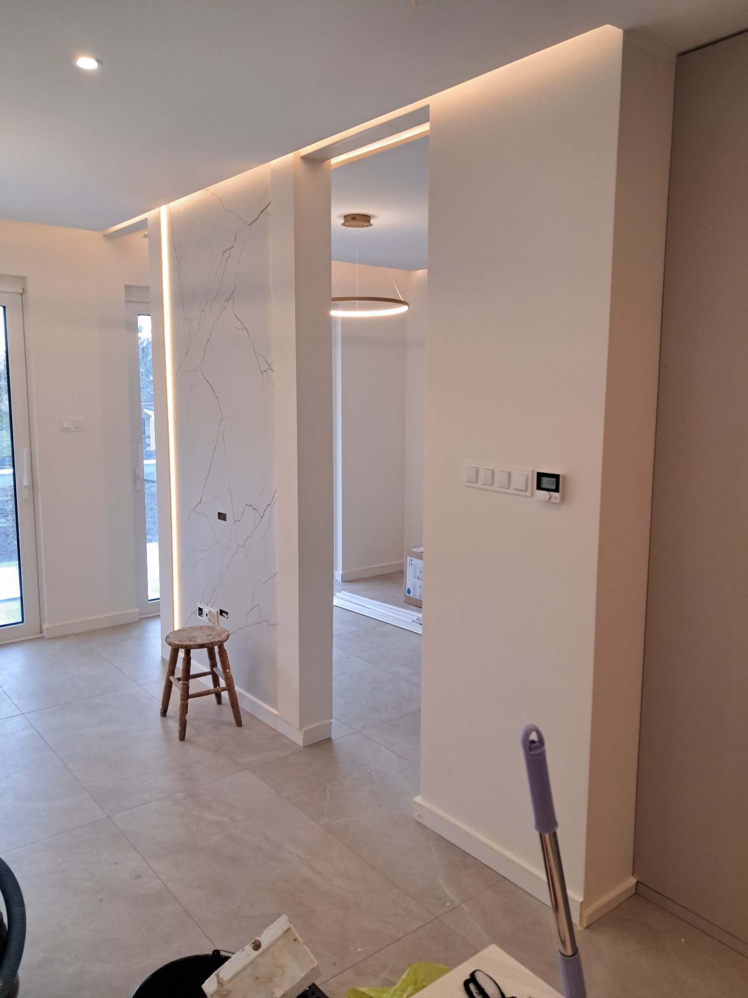 Nowoczesne wnętrze z podświetlanymi panelami imitującymi marmur i ledowym oświetleniem sufitowym. Widoczny fragment pomieszczenia z krzesłem i akcesoriami malarskimi.