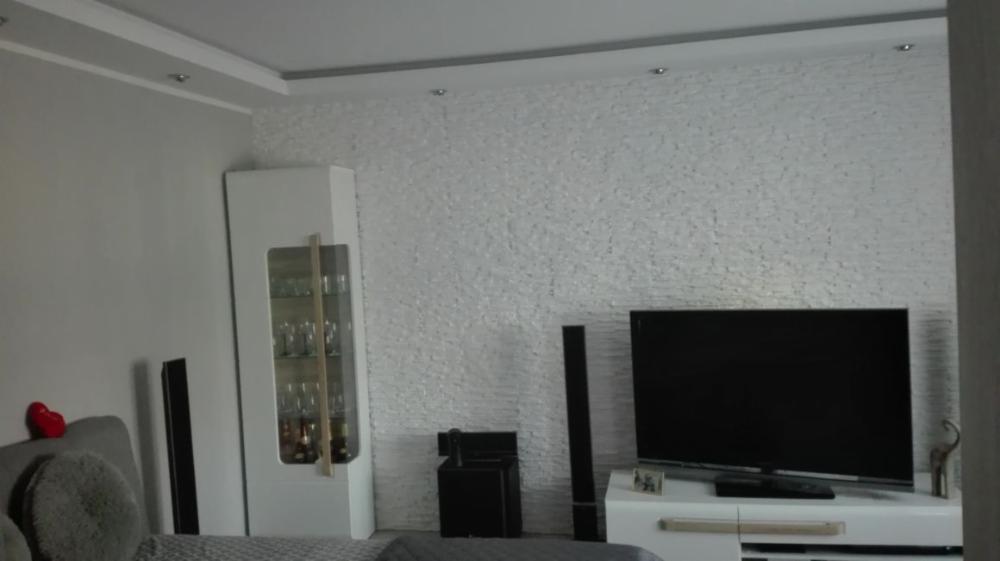 Jasne wnętrze salonu z dekoracyjną ścianą z białego kamienia, białymi meblami i podświetlanym sufitem, widok z perspektywy kanapy.