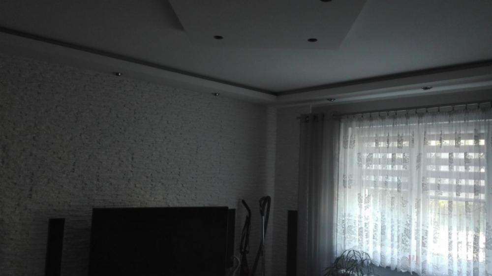 Wykończone wnętrze: ściana pokryta dekoracyjną białą cegłą, sufit podwieszany z oświetleniem punktowym i karnisz z zasłonami w oknie.