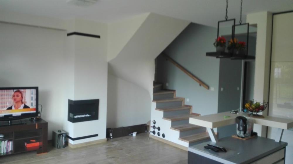 Jasne, nowoczesne wnętrze z kominkiem obudowanym w ścianie, schodami z drewnianymi stopniami i wnękami na wino, oraz wyspą kuchenną z dekoracyjną lampą wiszącą i misą owoców.