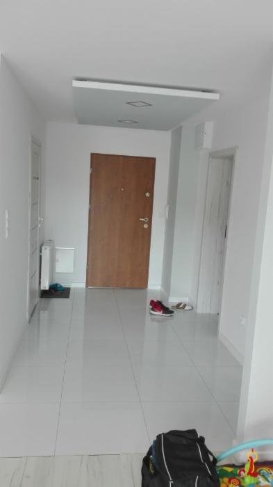 Jasne wnętrze przedpokoju z białymi ścianami i podłogą z płytek, drewniane drzwi wejściowe, podwieszany sufit z oświetleniem punktowym, widoczne buty i torba na podłodze, w tle drzwi do innego...