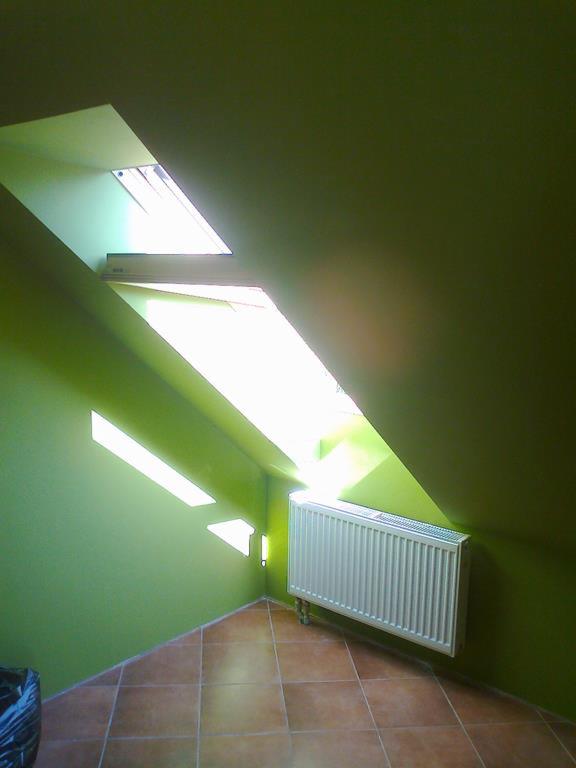 Pomieszczenie na poddaszu z zielonymi ścianami, brązowymi płytkami na podłodze, białym grzejnikiem i oknem dachowym w kształcie trapezu wpuszczającym jasne światło.