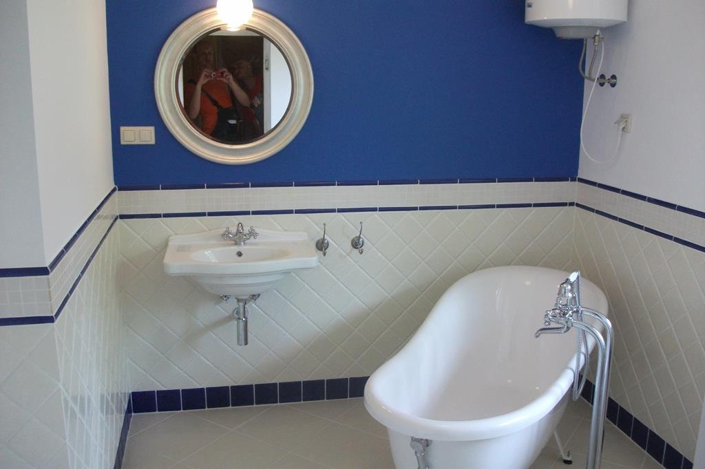 Biała łazienka z niebieskimi akcentami, owalne lustro z odbiciem osoby robiącej zdjęcie, bateria wannowa wolnostojąca, biała umywalka na ścianie, biało-niebieskie płytki ułożone w karo.