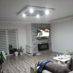 Jasne wnętrze salonu z panelami podłogowymi, ceglaną ścianą z kominkiem i telewizorem, oraz oświetleniem sufitowym w kształcie kwadratu z wbudowanymi lampami