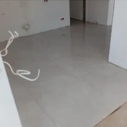 Jasne, kwadratowe płytki podłogowe z widocznymi fugami, ułożone w pustym pomieszczeniu w trakcie remontu, z wystającymi przewodami elektrycznymi przy ścianie.