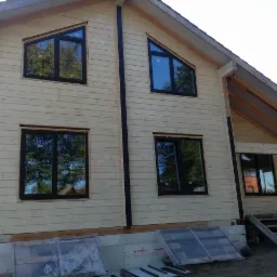 Surowy dom z drewnianych bali z czarnymi ramami okiennymi i folią ochronną na oknach na parterze.