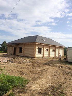 Surowy, parterowy dom z drewnianych bali z ciemnoszarą dachówką, w trakcie budowy, widoczny na tle błękitnego nieba i przygotowanego terenu.